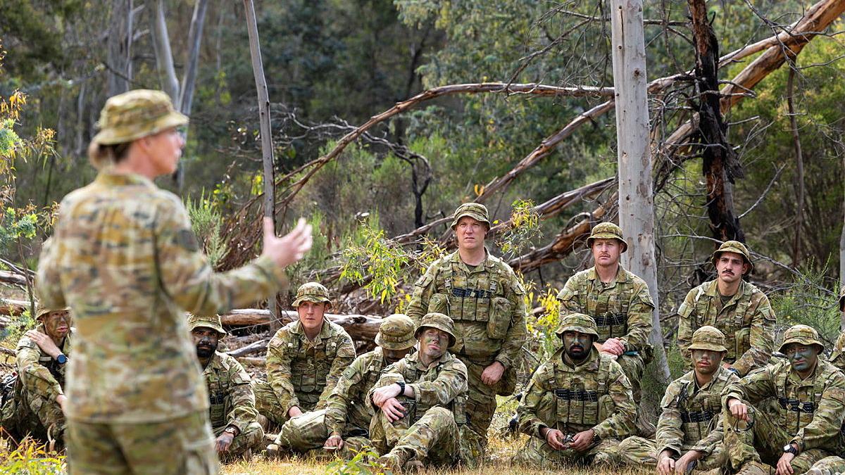 People in Army uniform sit on grass while a woman in uniform speaks to the group.