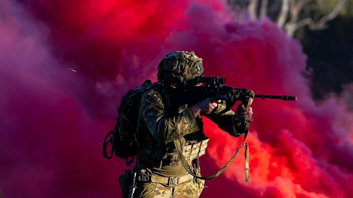 A person in Army uniform shoots a rifle, with the shell flying out behind them, against a backdrop of pink smoke.