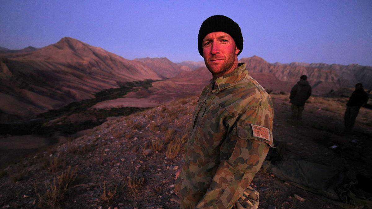 A man in uniform and wearing a dark beanie stands in a rugged mountainous landscape beneath a purple sky.