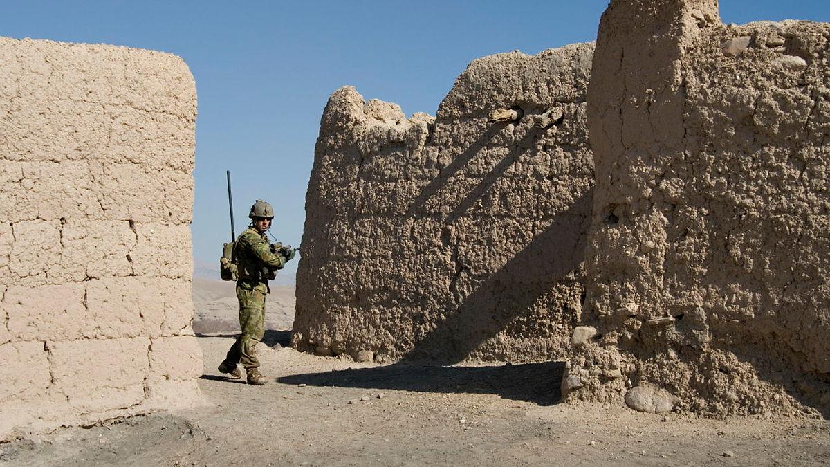 An Army soldier on patrol on a desert with dirt walls either side of him.