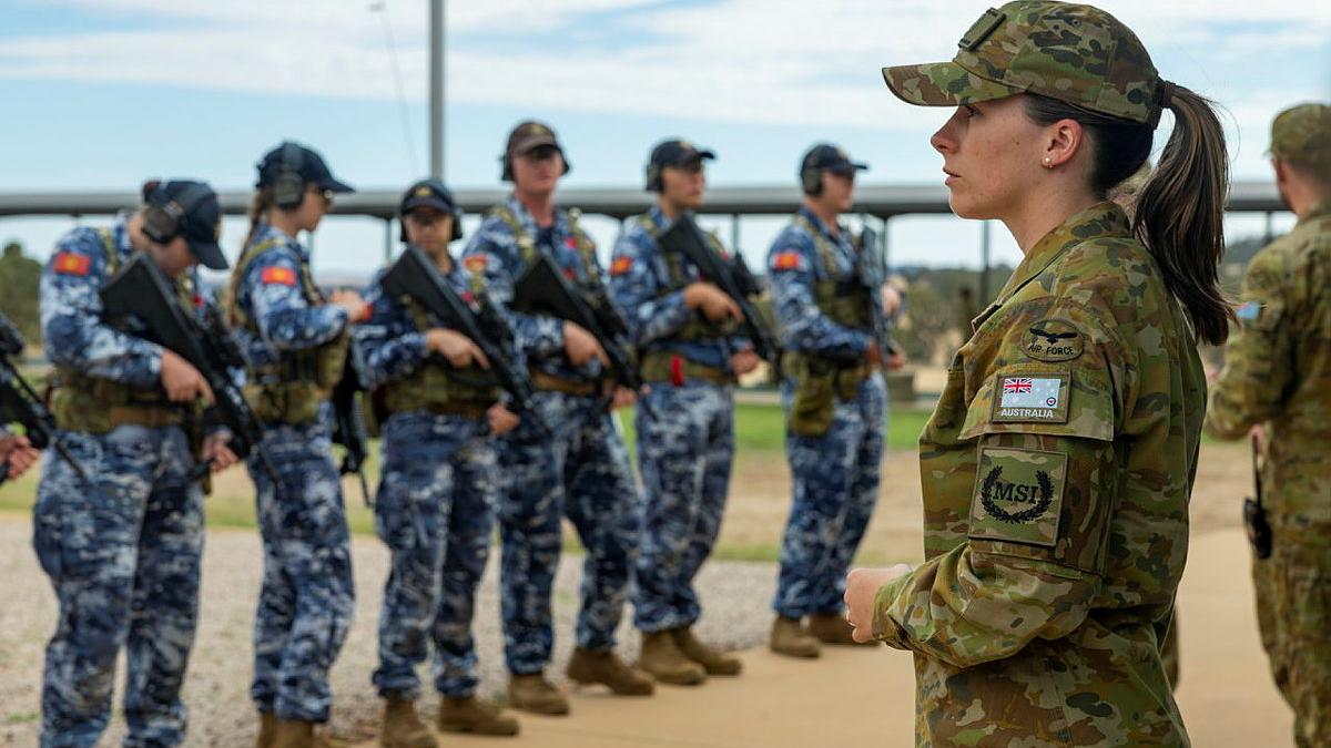 Two military personnel in green camo uniform survey a row of armed military personnel in blue camo uniform.