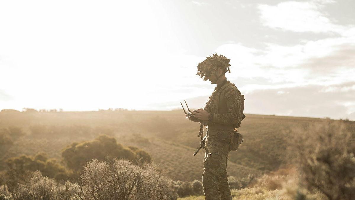 A man in camouflage uniform stands in a grassy open area holding a controller, with scattered trees and shrubs in the background.