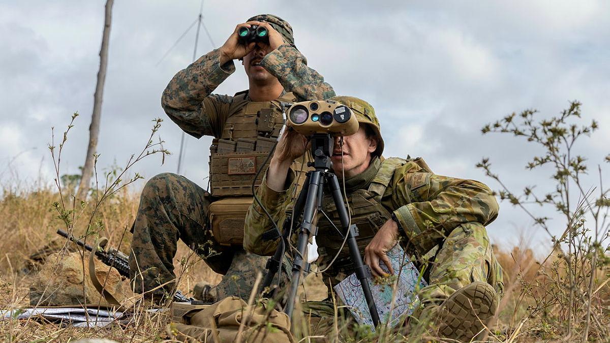 Two soldiers in different uniforms look through binoculars in an outback location.