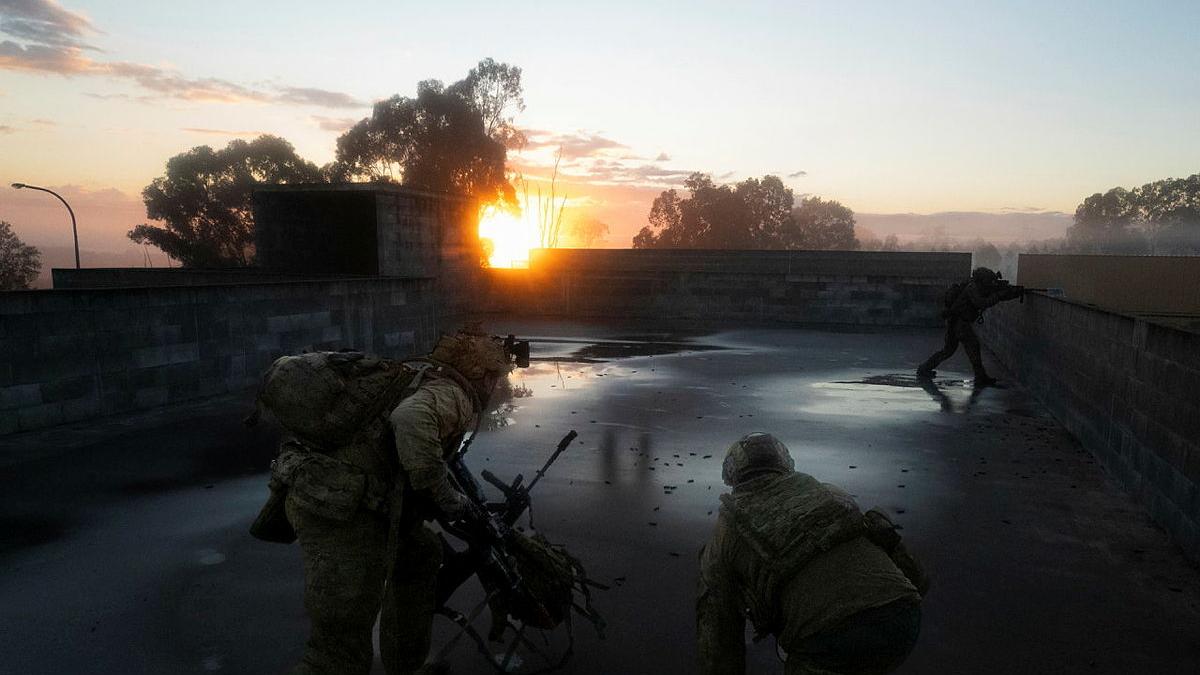 Three men in uniform occupy positions on a wet concrete surface, with one aiming a weapon over a concrete wall as the sun rises in the distance.