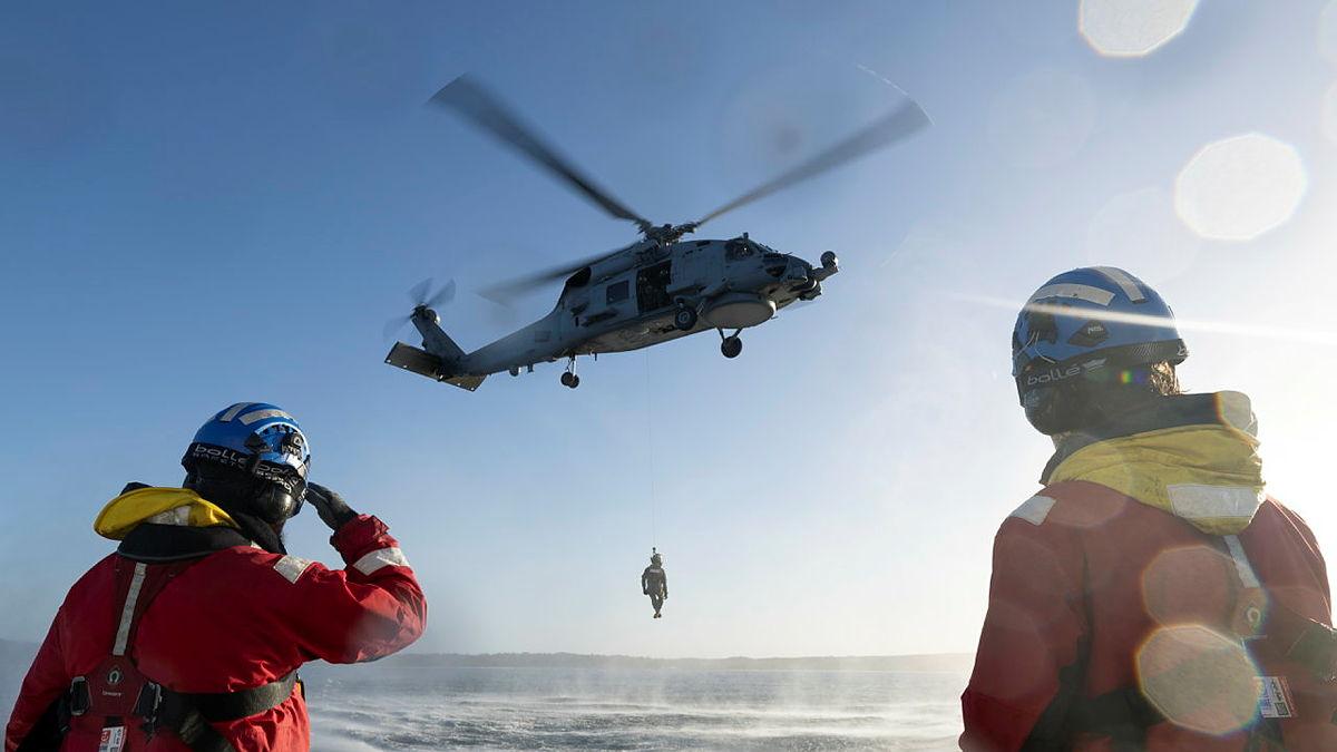 A helicopter hovers over water with a person suspended from a winch underneath while two people in helmets and orange jackets watch.