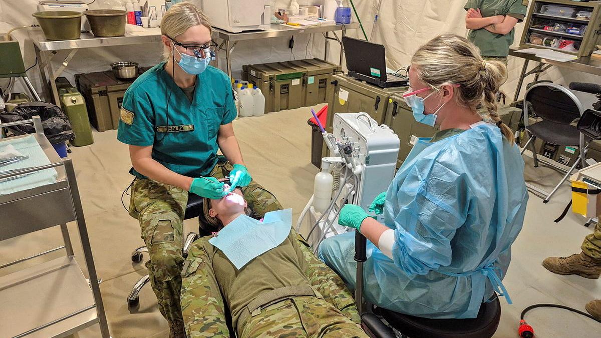 Soldiers in scrubs perform dental work on another solider.