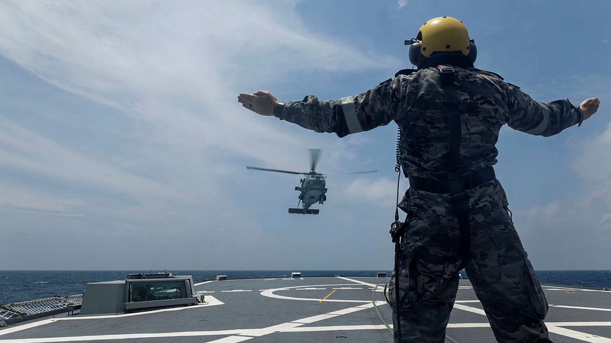 A military man wearing a helmet signals with his arms to an incoming helicopter, while onboard a ship at sea.