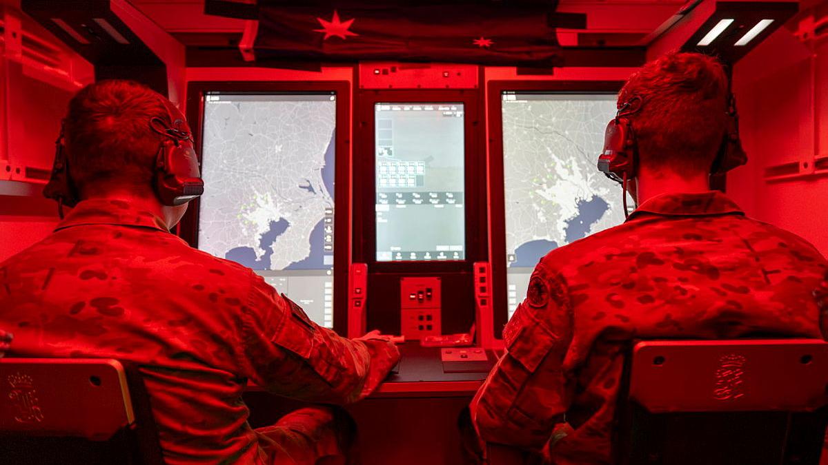 Two people in uniform sit looking at screens in a small room with red lighting.