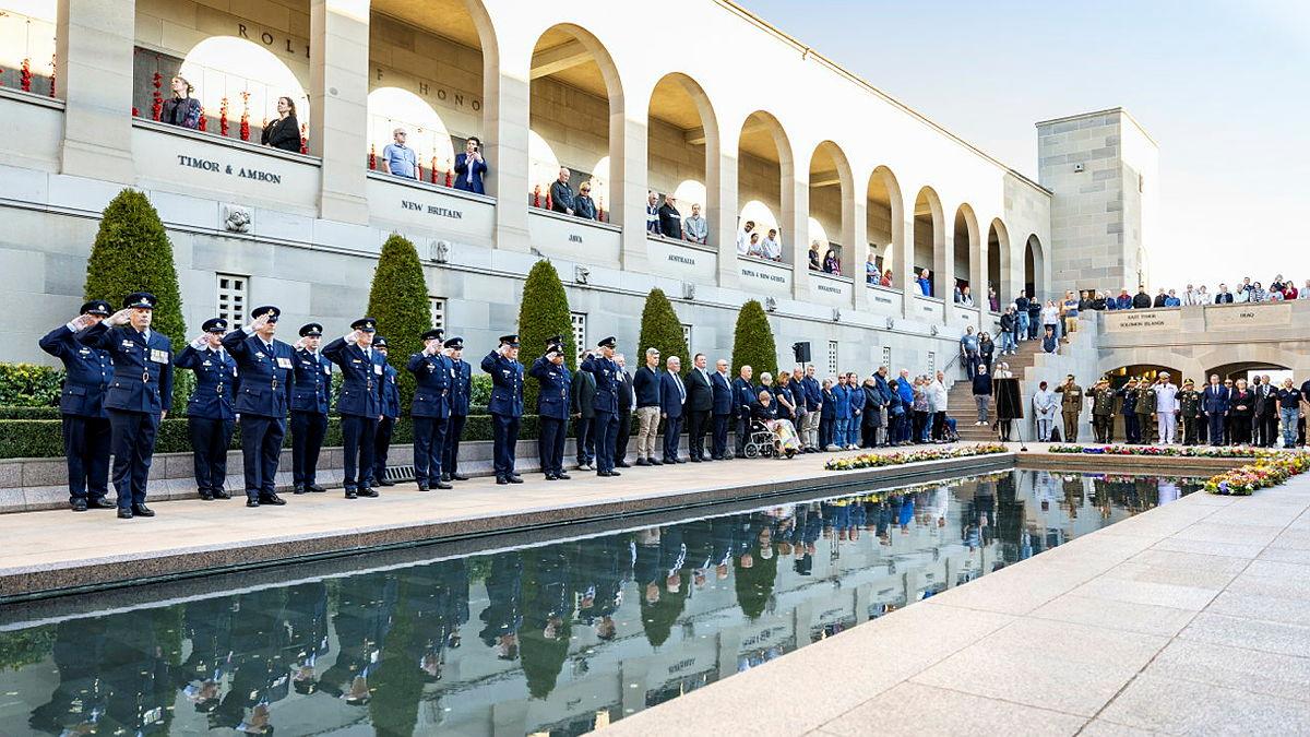 People in uniform salute beside a long reflective pool as people watch from balconies above.