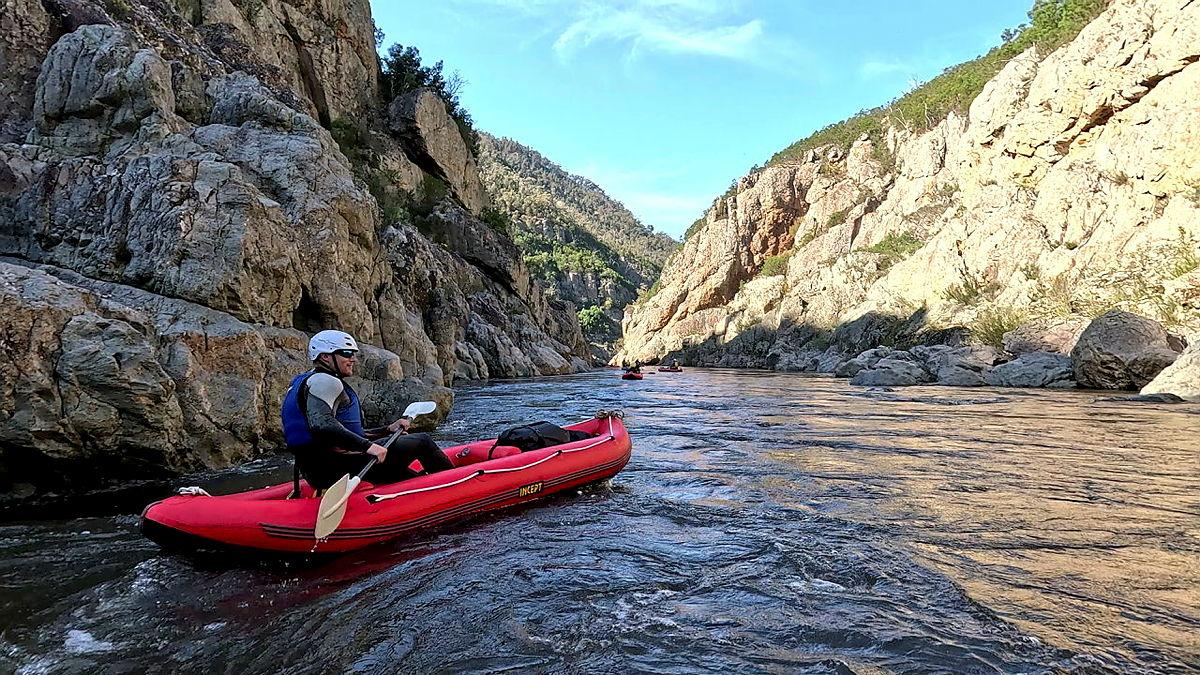 A person wearing a helmet and in a kayak floats on a river flanked by high rocky terrain.