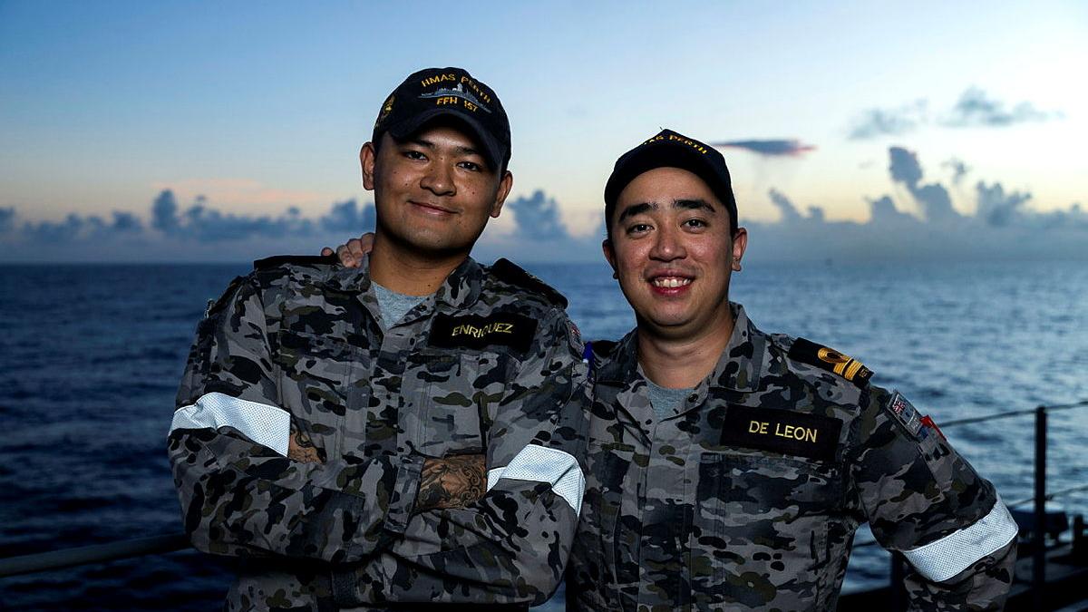 Two men in Navy uniform stand side by side on the deck of a ship with the sea behind them.