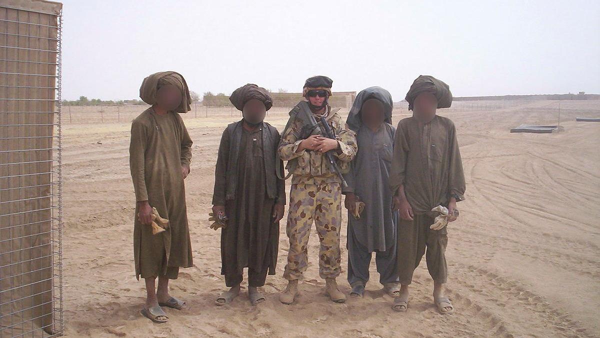 A military man stands with four civilians, whose faces have been blurred out, in the desert.