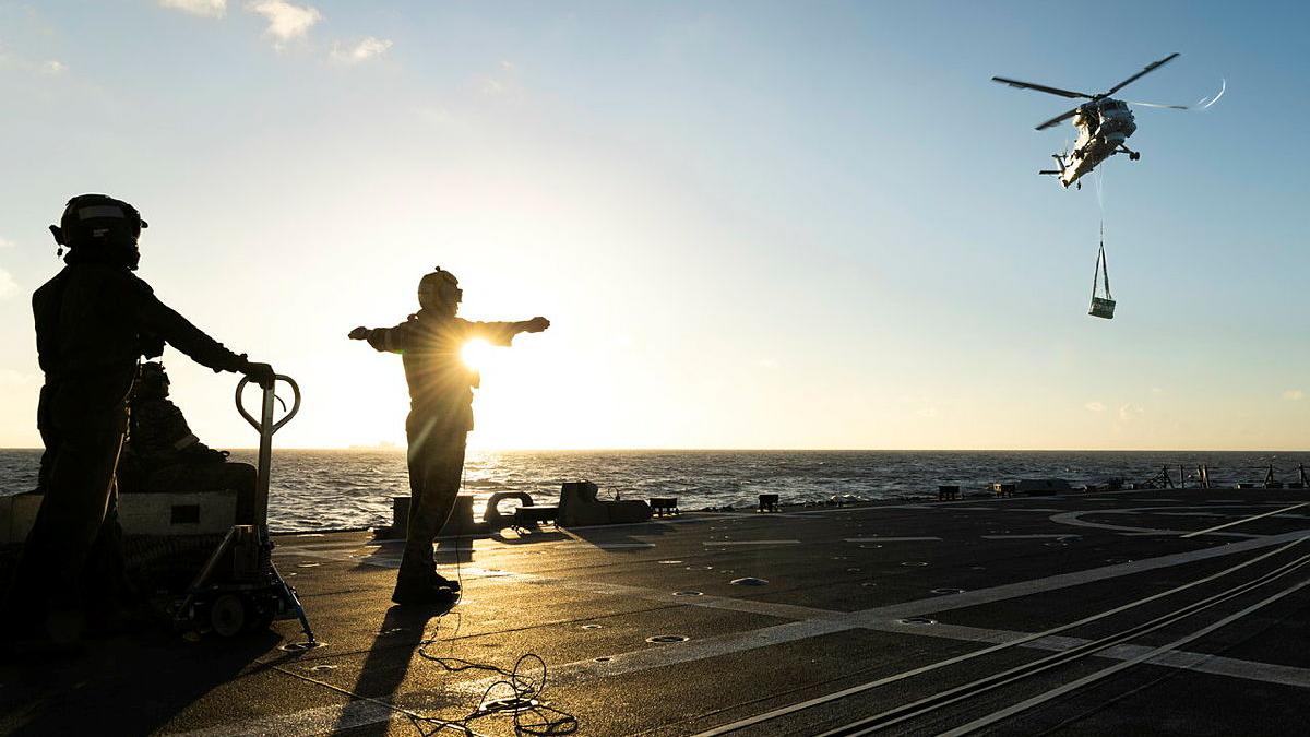 Three people on the flight deck of a ship are silhouetted by the sun as a helicopter hoists a large box over the sea towards the deck.