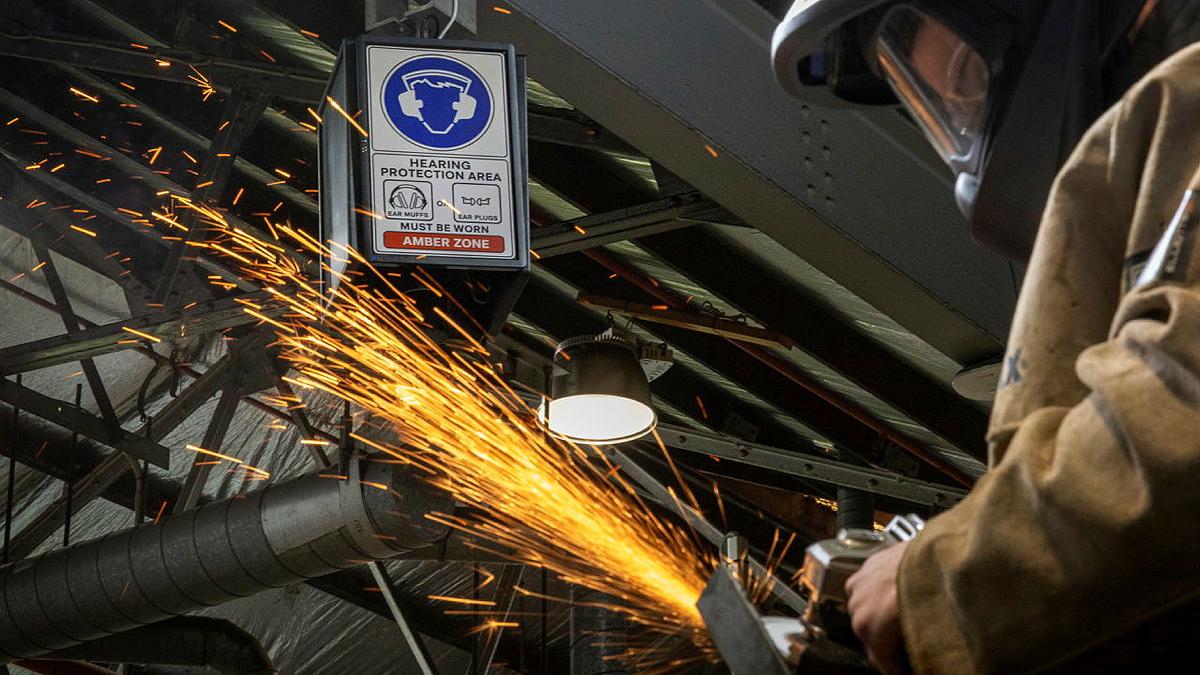 A person in safety protection gear works with metal, causing sparks to fly. A noise protection sign hangs nearby.