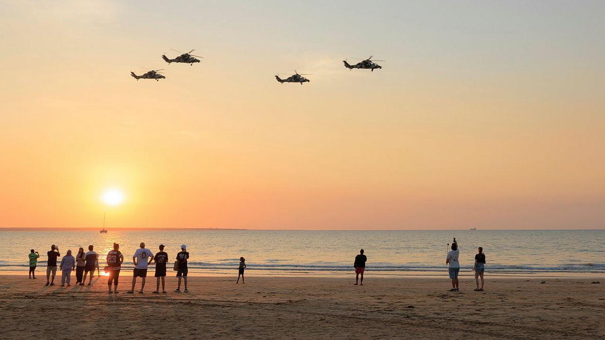 From the beach, civilians look up at four helicopters flying through a sunset sky.