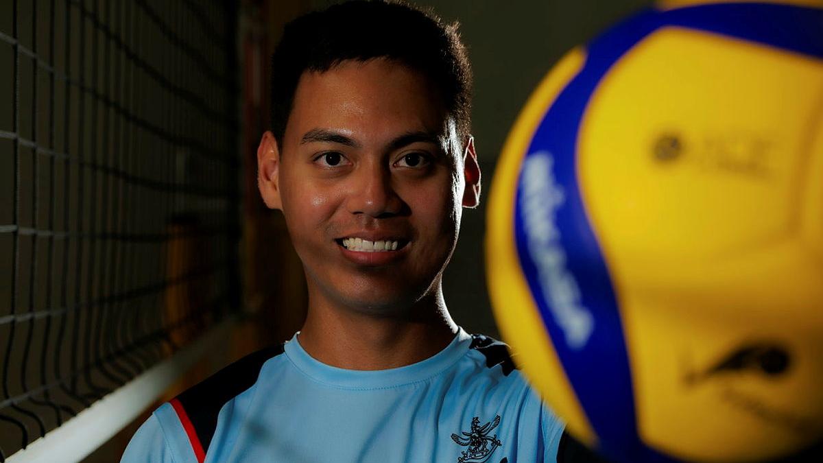 A man holds up a volleyball to the camera while standing next to a volleyball net.