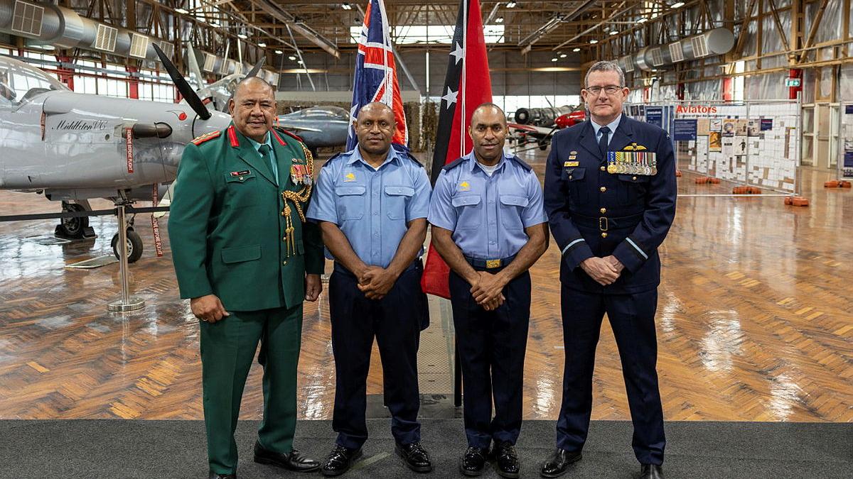 Four military personnel stand in a warehouse with stationary aircraft and two national flags.