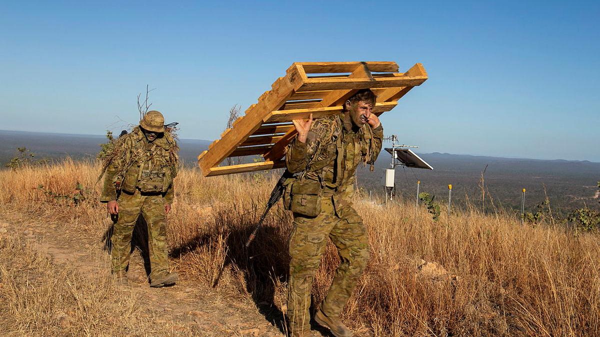 Two soldiers on a grassy hilltop, one of which is carrying a wooden crate.