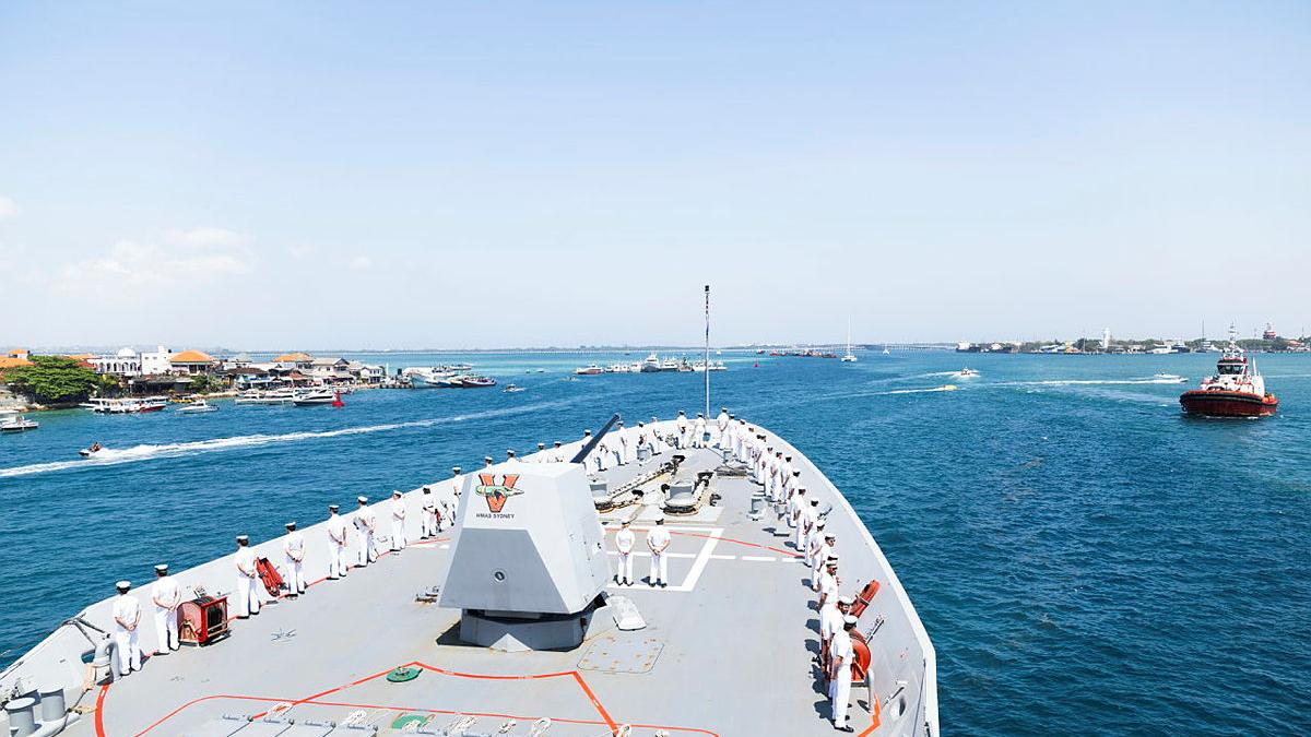 Sailors line the deck of a ship as it sails into a port.