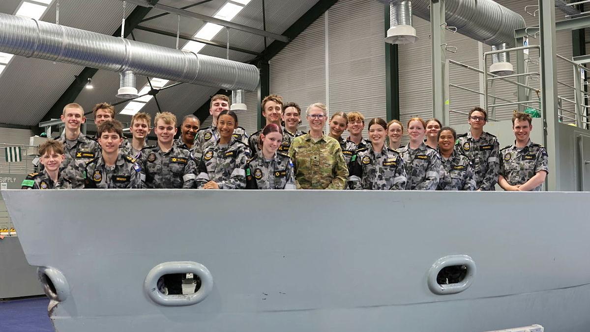 A group of people in Navy uniform stand in a small-scale replica of a ship’s bow inside a warehouse.