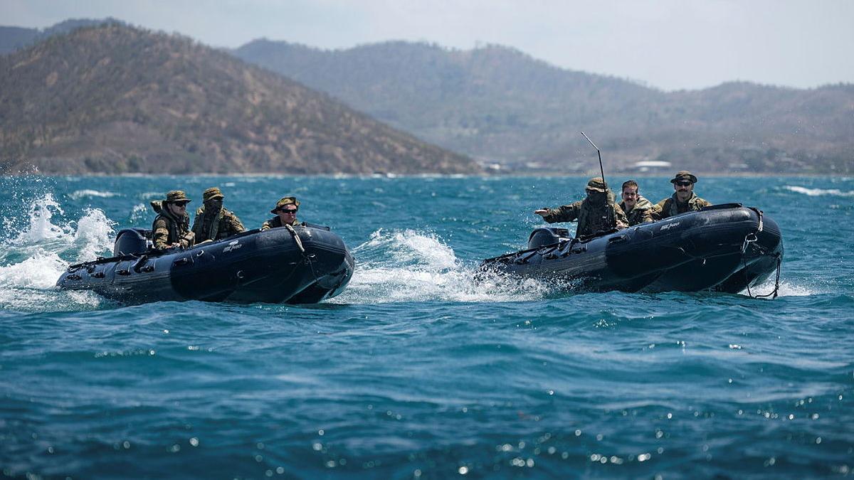 Two small boats carrying three people in uniform each travel through the water with a mountainous landscape behind them.