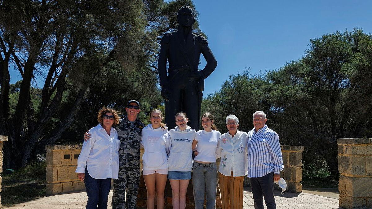 A man in a Navy uniform stands with a group of people in plain clothes in front of a large statue of a person.