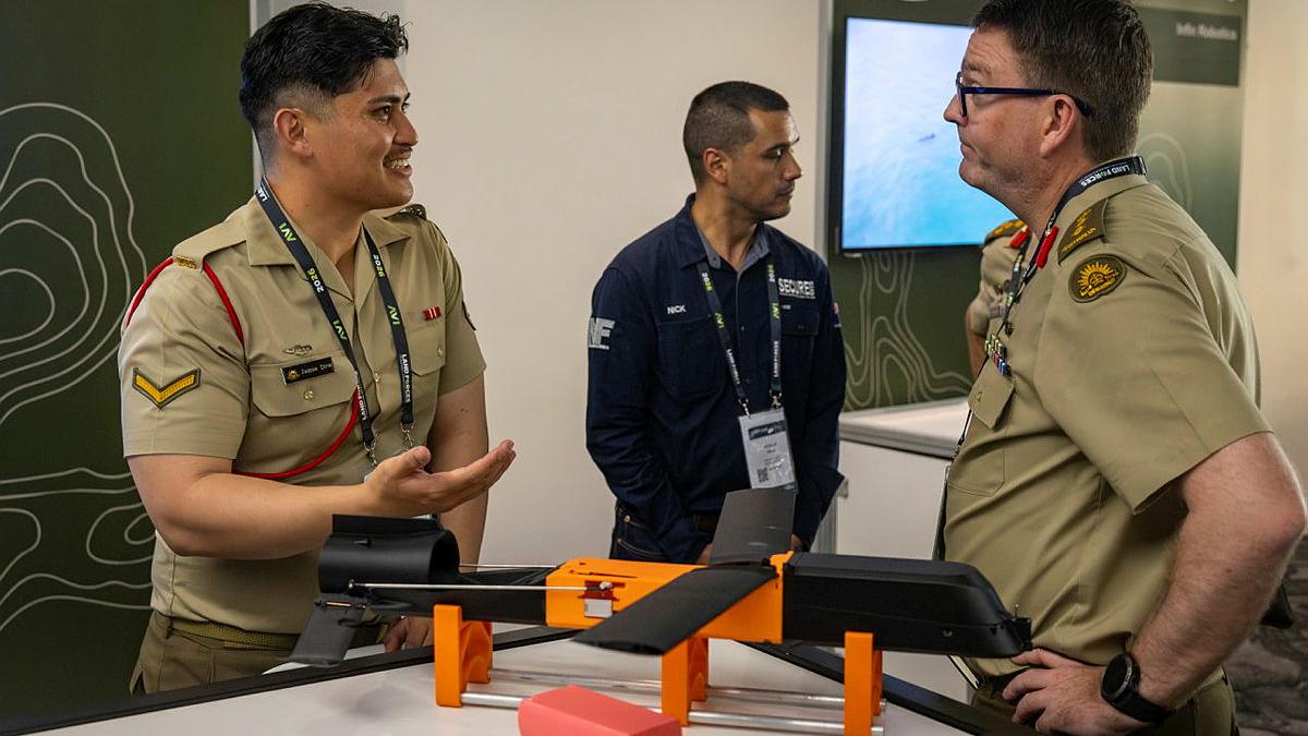 An orange and black device sits on a table between two men in Army uniform who are talking.
