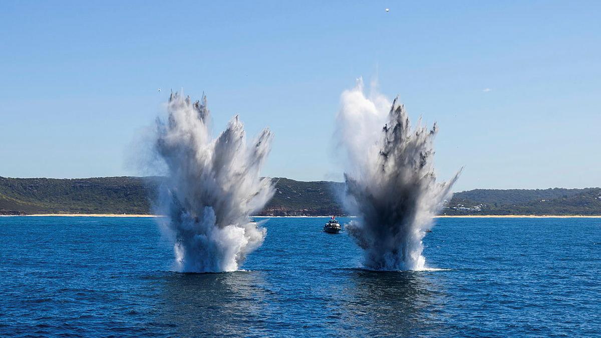 Two explosions of water side by side in a bay, a boat and shoreline in the distance.