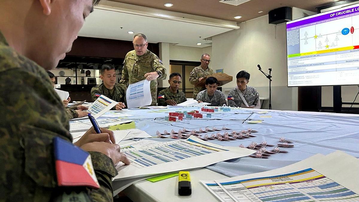 People in uniform sit around a large table looking at sheets of paper, while a large projector screen shows a slide in front of the table.