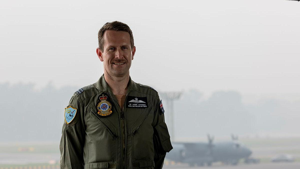 A military man stands before a window looking out at a foggy day on an airfield tarmac.