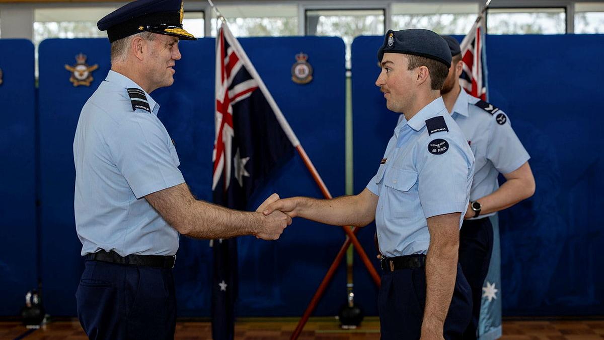 Two male military members shake hands in front of an Australian flag.