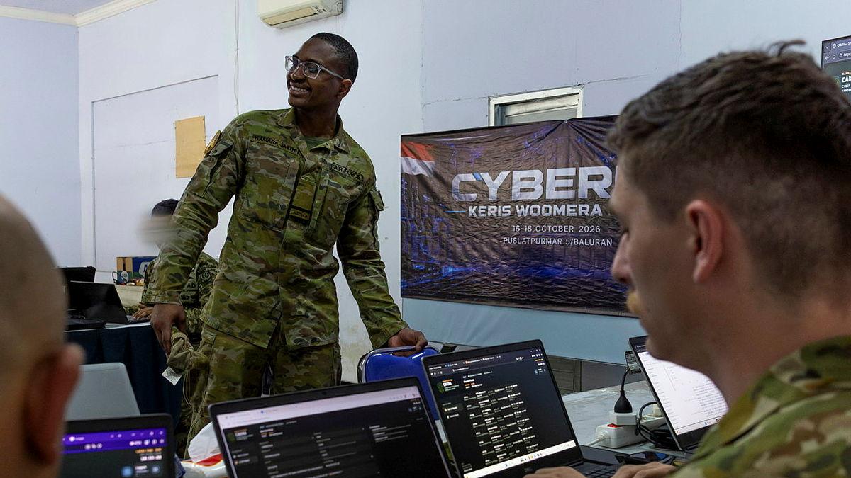 A man in military uniform stand in front of a class on military members working on laptops.