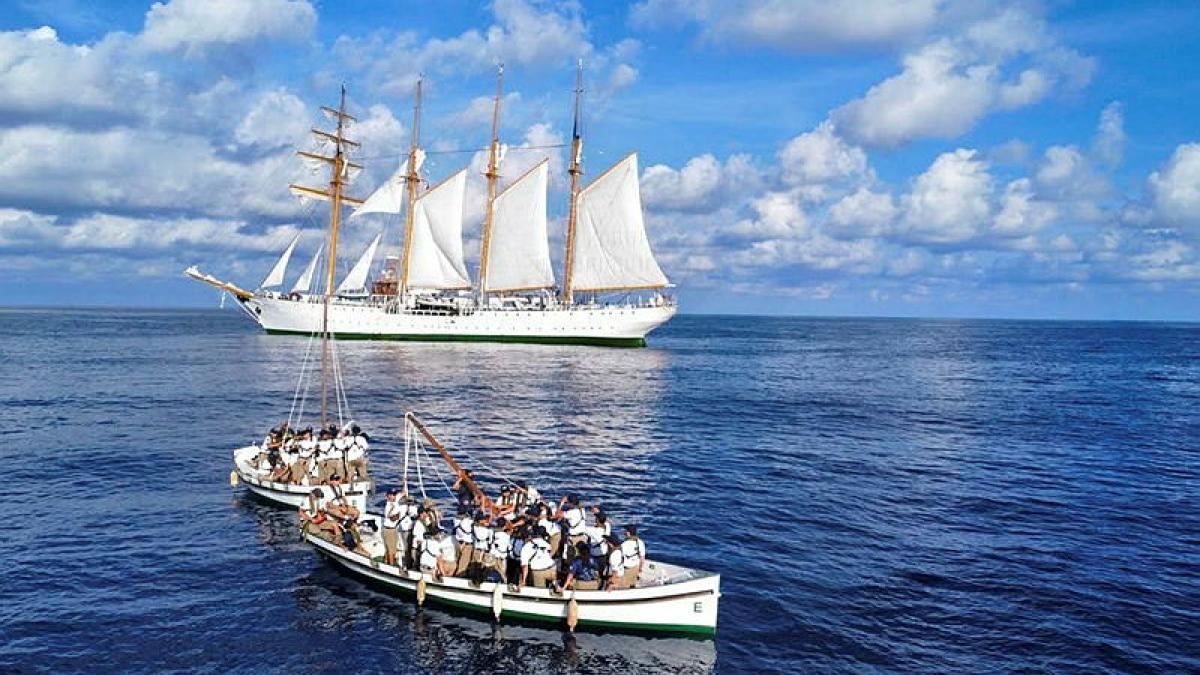 Two small sail boats, each filled with people, are in the ocean a short distance away from a tall ship.