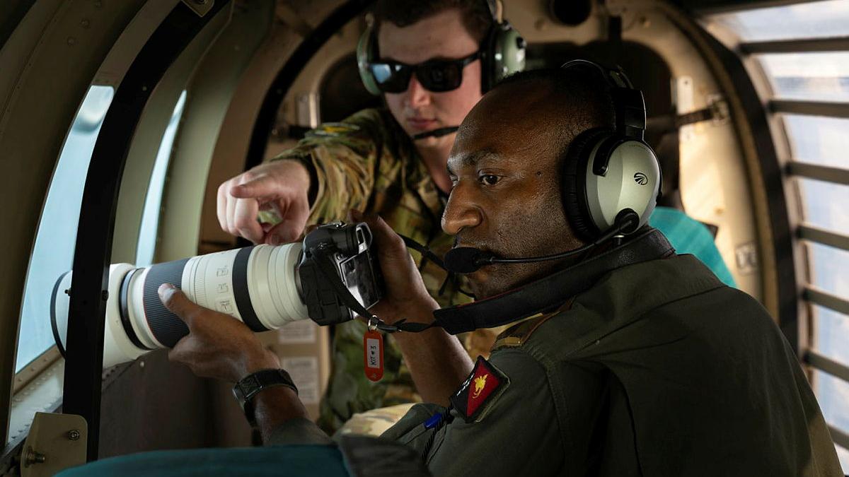 A military man instructs another military man with a camera, while both are inside an aircraft.