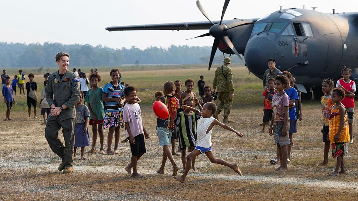 A military man watches a child kick a football. Other children and an aircraft are nearby.