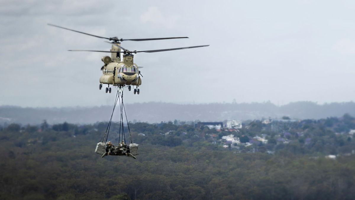A helicopter flies over tree tops with a large object suspended beneath it. 
