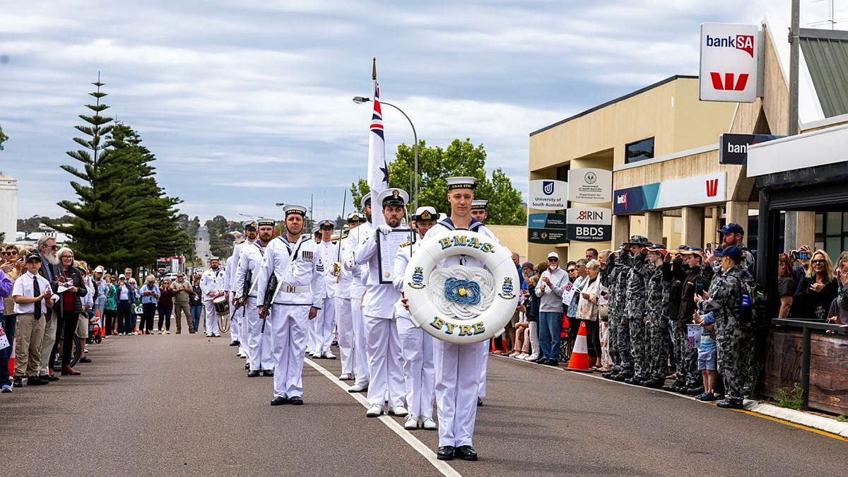 A group of Navy personnel in white uniforms march down a street with people watching from the side..