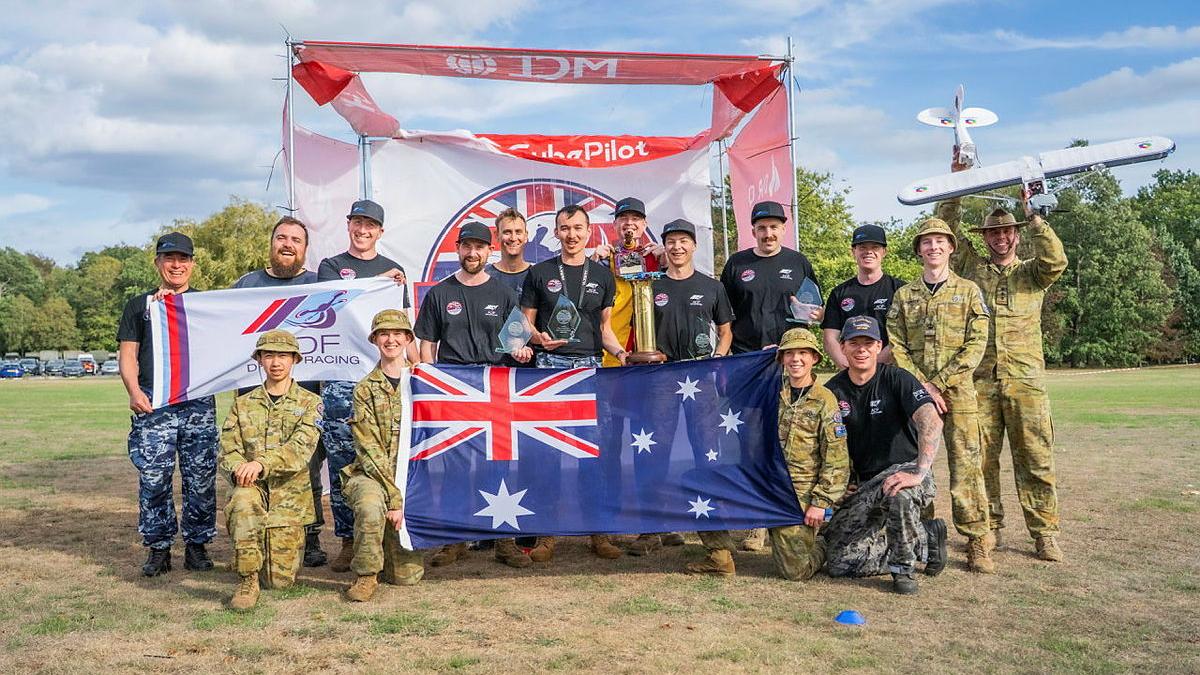 A group of military personnel stand with an Australian flag, a trophy, a drone and a drone racing banner. 