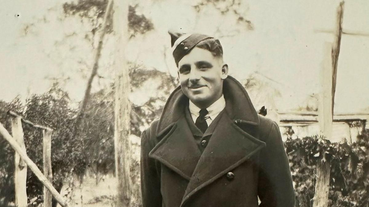 A young man wearing a coat and uniform cap stands outdoors in a black-and-white photograph.